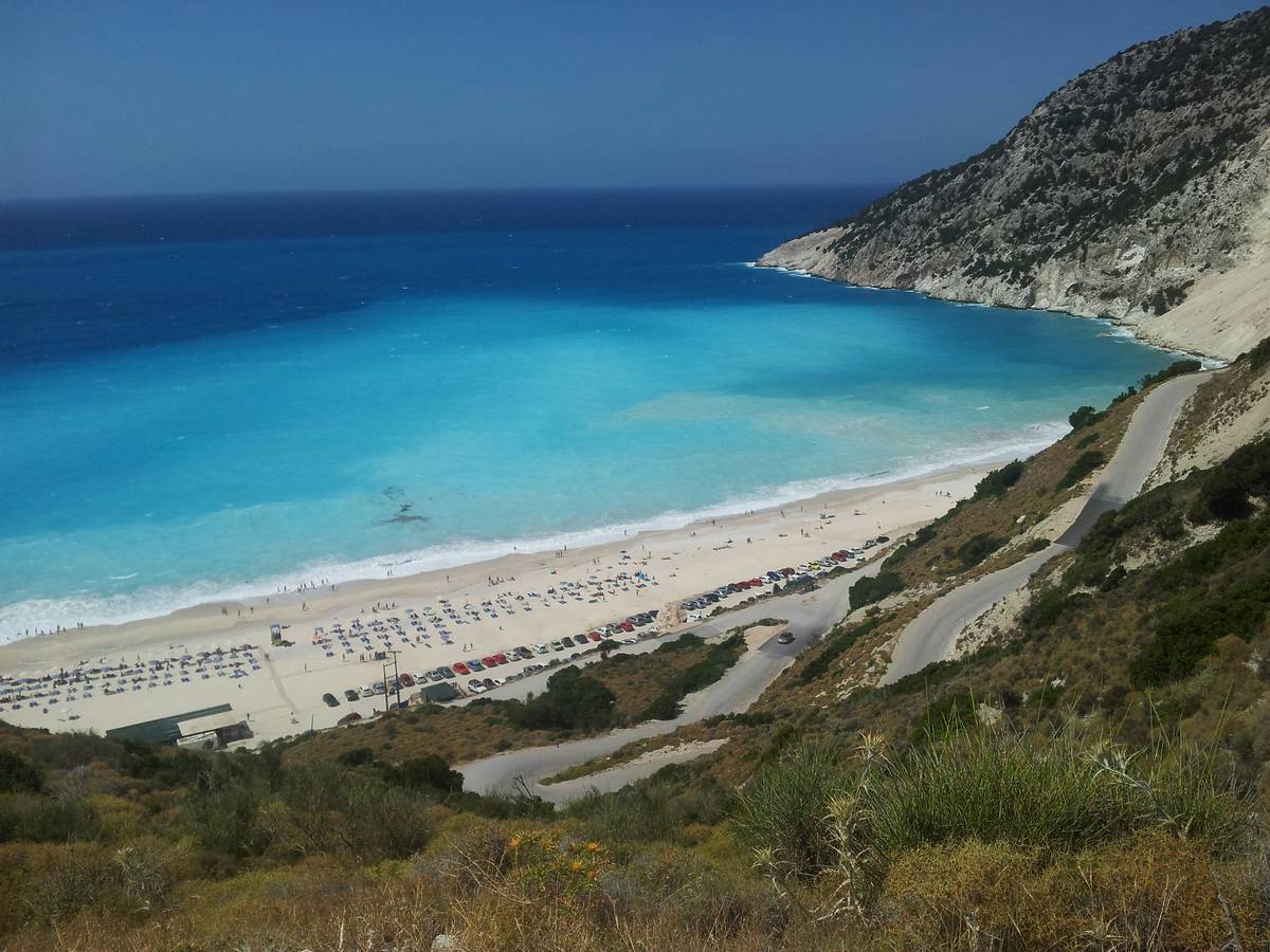 Turquoise sea with a white sandy beach with lots of chairs on seen from a distance. A green hill is seen in the foreground.