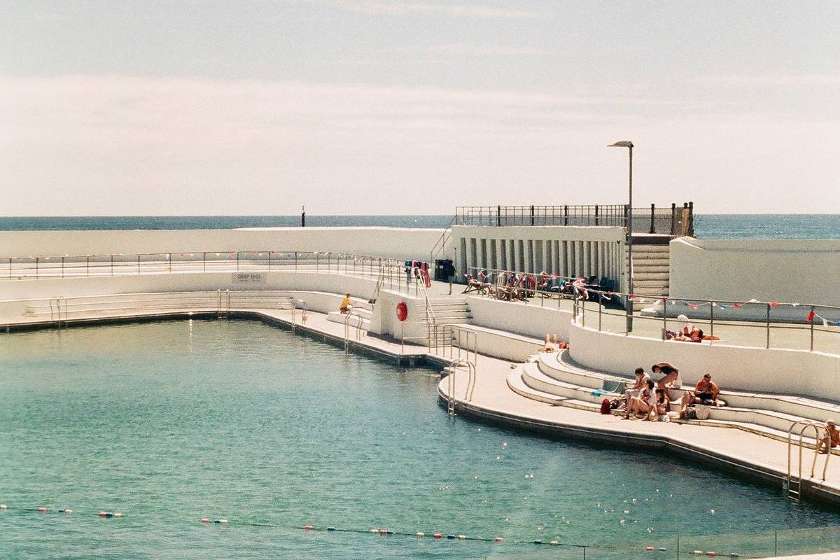An art-deco style lido with people lounging on the stairs surrounding the pool on a sunny day.