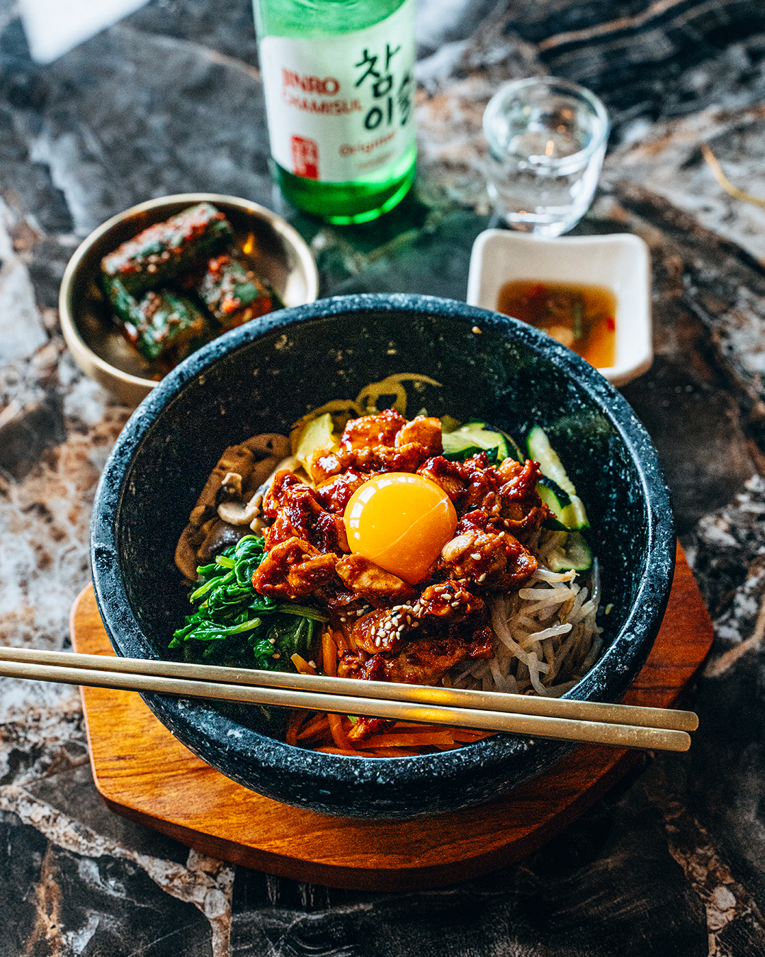 a bowl of korean food with chopsticks placed on the rim, surrrounded by sauces, side dishes, and drinks