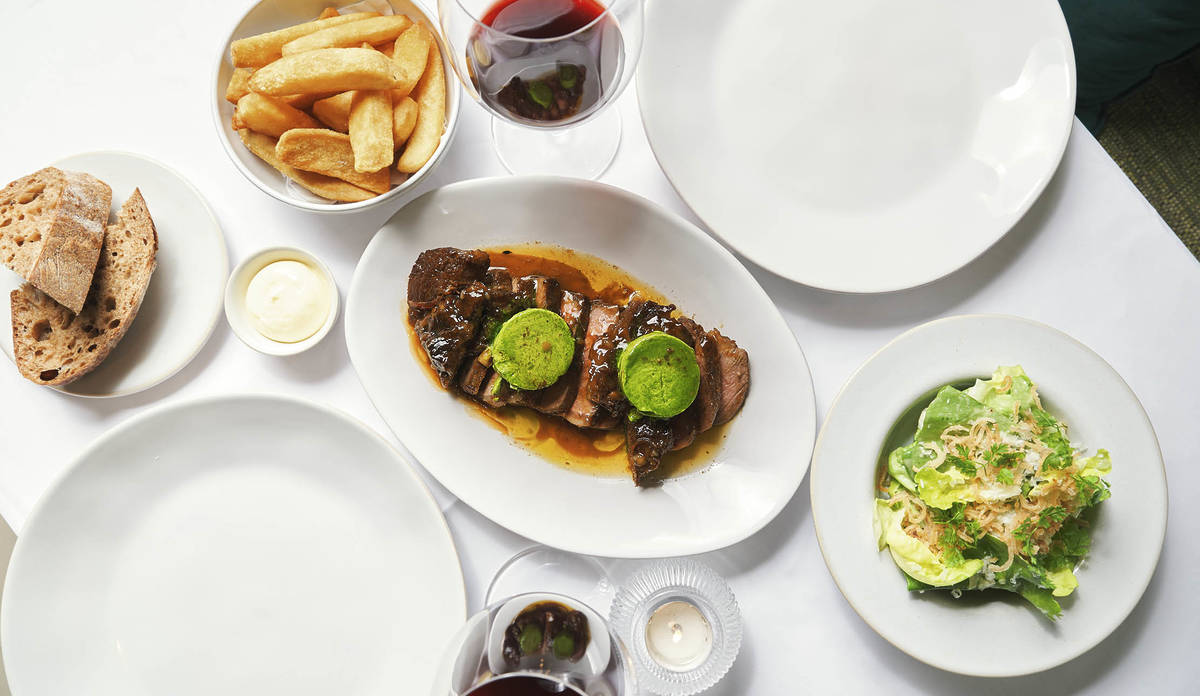 a platter of steak in the middle of a table, also laid with plates, salad, chips, and glasses of wine