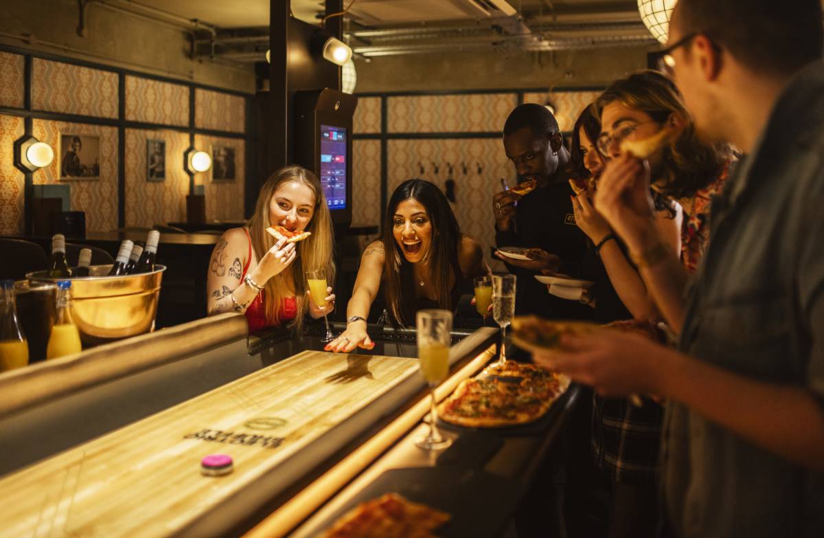 Woman playing shuffleboard while friens watch, drink, and eat pizza.
