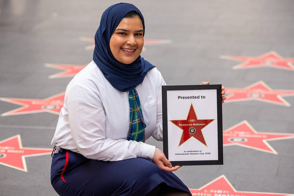 Rezwana Rahman ajoelhada ao lado de uma estrela, enquanto segura uma estrela emoldurada, numa trilha de estrelas que celebra as mulheres no setor ferroviário