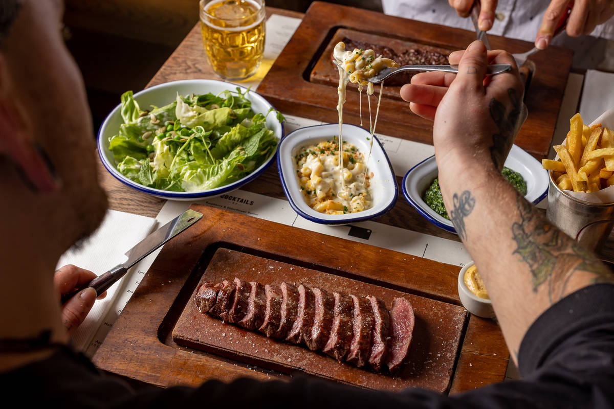 someone digging into a portion of mac and cheese from a table covered in steaks, drinks, and side dishes