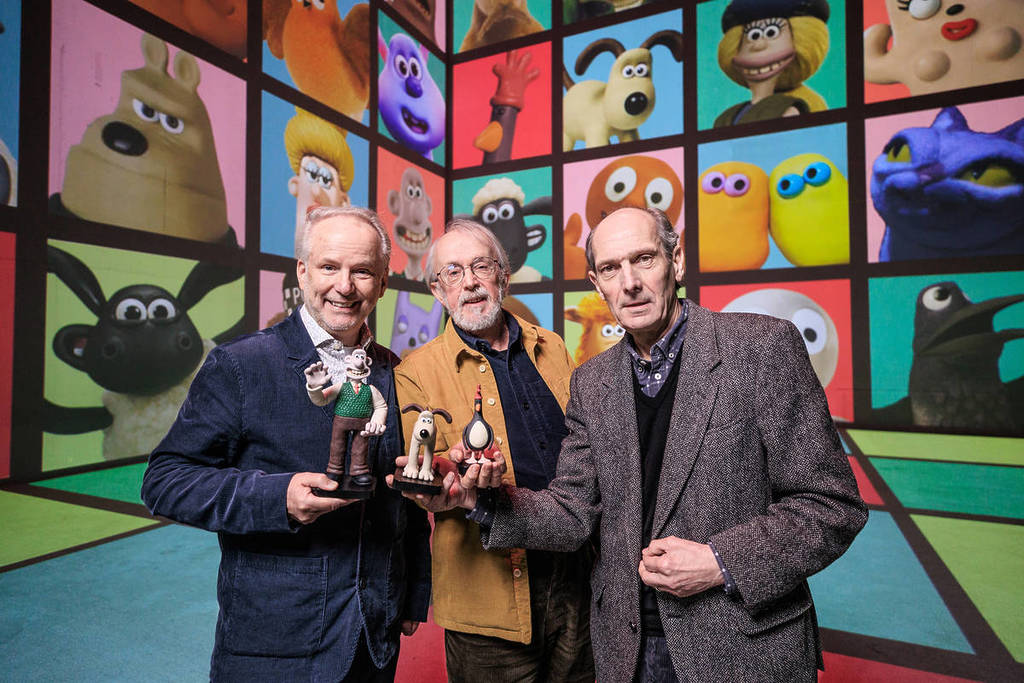 Die Schöpfer und Mitbegründer von Aardman, Nick Park, Peter Lord und David Sproxton, halten Tonfiguren vor einem farbenfrohen Bildschirm voller ihrer Claymation-Figuren