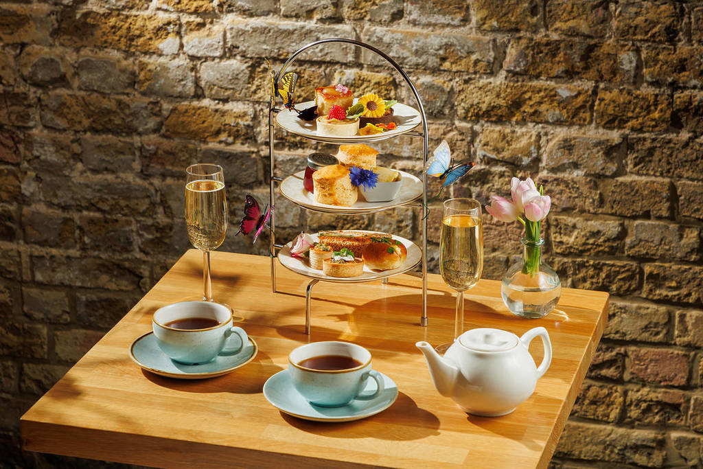 uma mesa posta para o chá da tarde com canecas de chá, copos de espumante e uma bandeja de três andares repleta de scones, sanduíches e doces, decorada com borboletas e flores