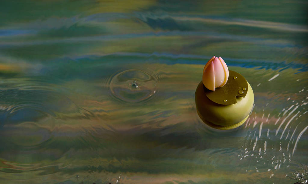 um doce inspirado na arte, em tons de verde e castanho, coberto com uma cobertura rosa semelhante a um lírio, apresentado contra um fundo inspirado em Monet, com água e cor