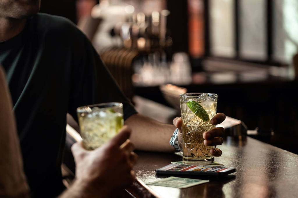 uma foto de duas bebidas num pub, uma erguida na mão de alguém, a outra no balcão com uma mão à volta dela