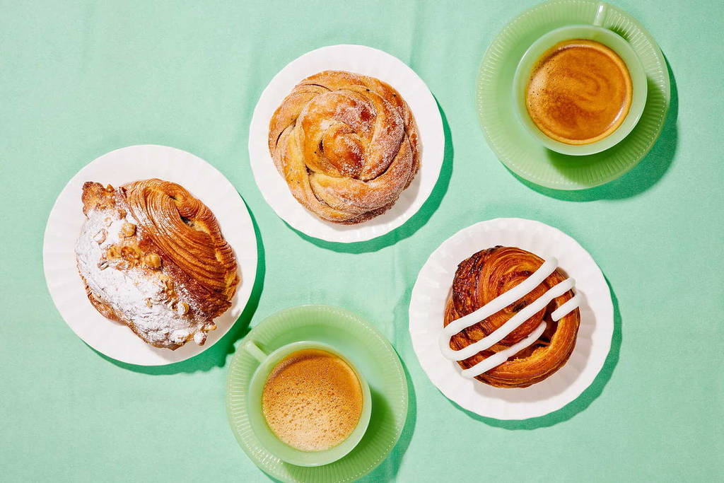 Eine Vogelperspektive auf eine Auswahl an süßem Gebäck auf kleinen Tellern, die auf einem grünen Tisch um zwei Tassen Kaffee in grünen Tassen angeordnet sind