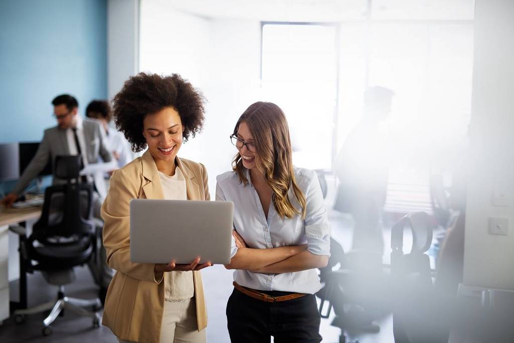 two business women working together in office; both are standing and one is holding a laptop open