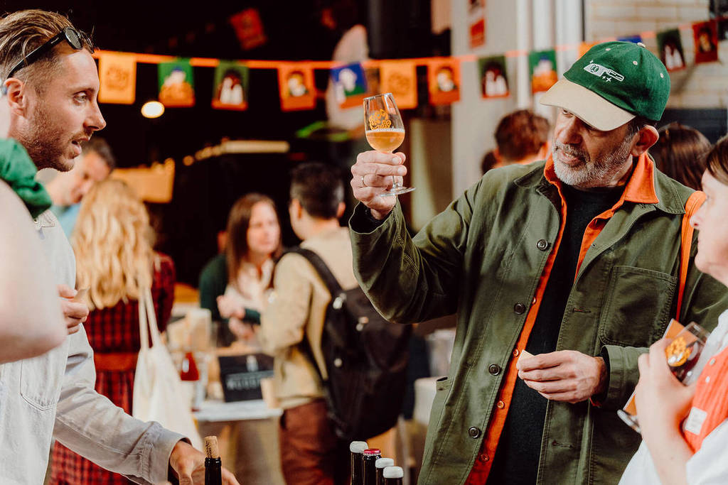 someone holding a glass of wine up to look at the colour, at a wine fesstival, as they talk to the wine producer and a friend