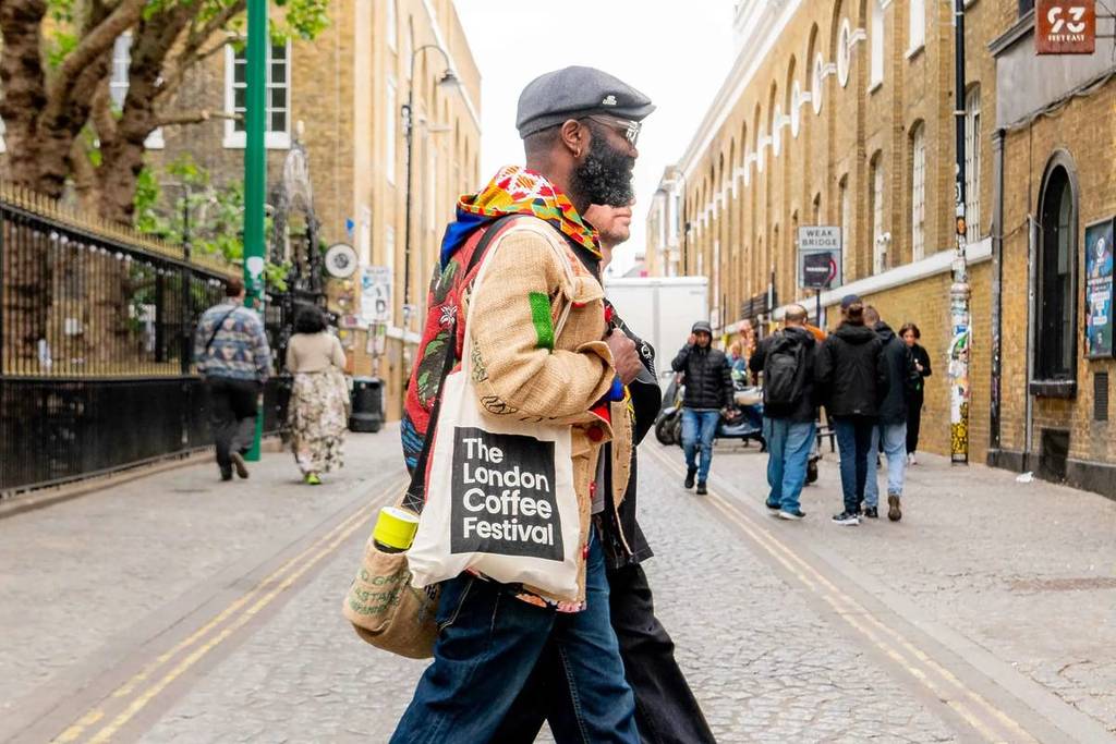 someone walking through the street in a bright scarf, cosy jumper, and cap, with a london coffee festival tote bag