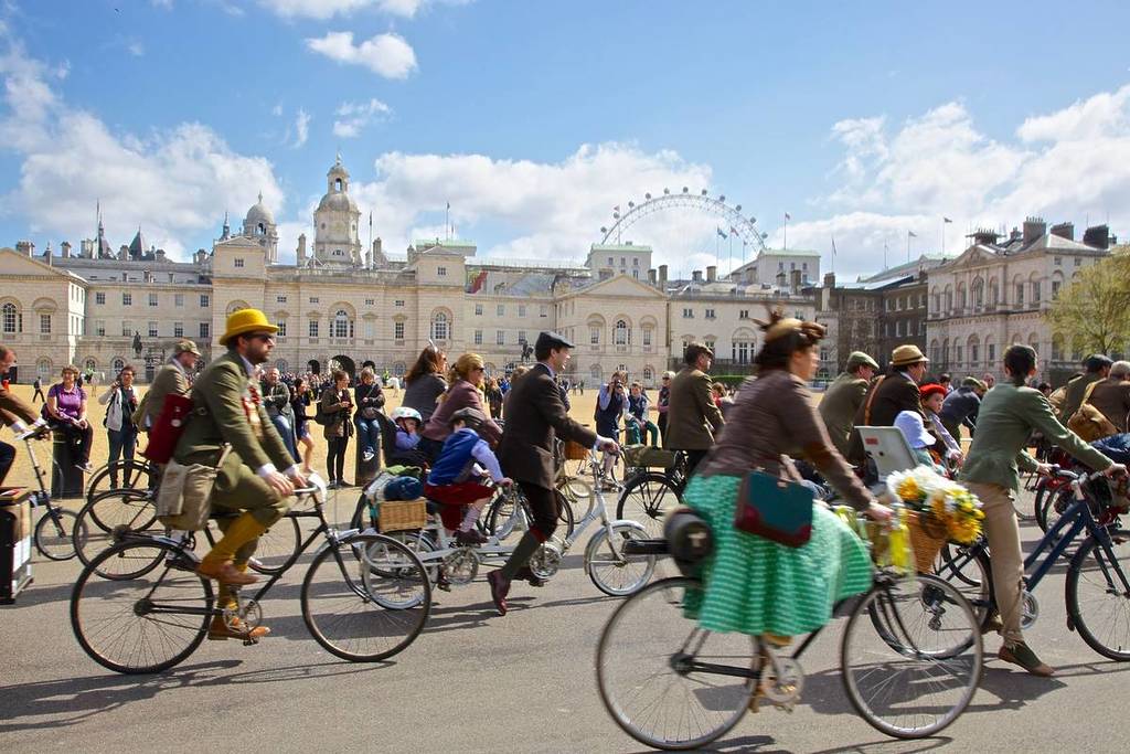 Menschen in Anzügen, schicken Hüten und eleganten Kleidern fahren mit dem Fahrrad durch London, das London Eye ist im Hintergrund zu sehen