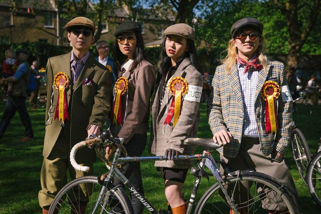 Four people dressed in tweed and flat caps stand behind a bike, wearing rosetres. 