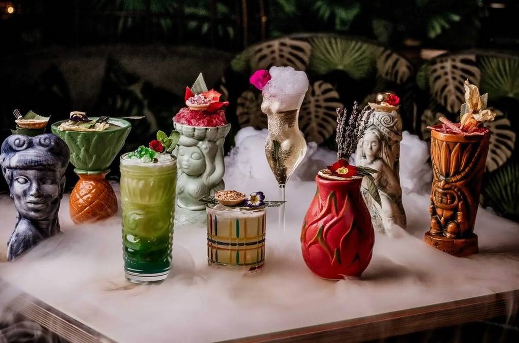 Eine Auswahl an Tiki-Cocktails in speziellen Gläsern und Bechern, umgeben von wirbelndem Nebel aus Trockeneis