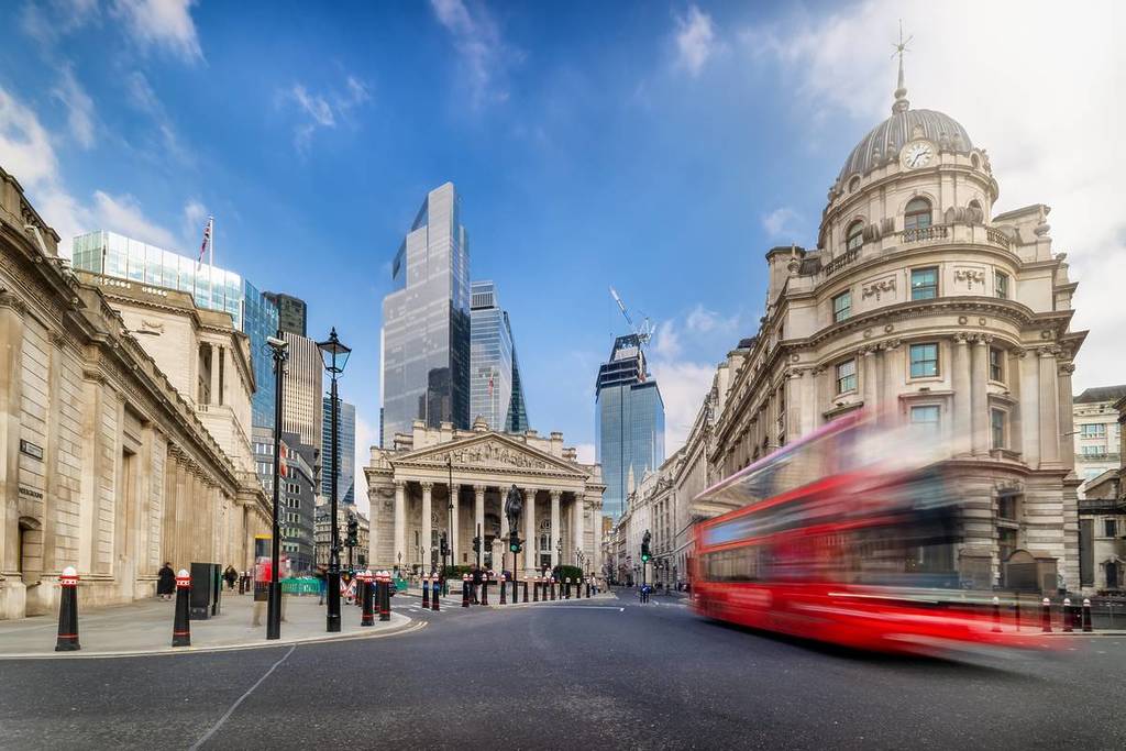 Langzeitbelichtung der City of London mit Straßenverkehr, darunter ein verschwommen vorbeifahrender roter Bus und die modernen Bürohochhäuser im Hintergrund