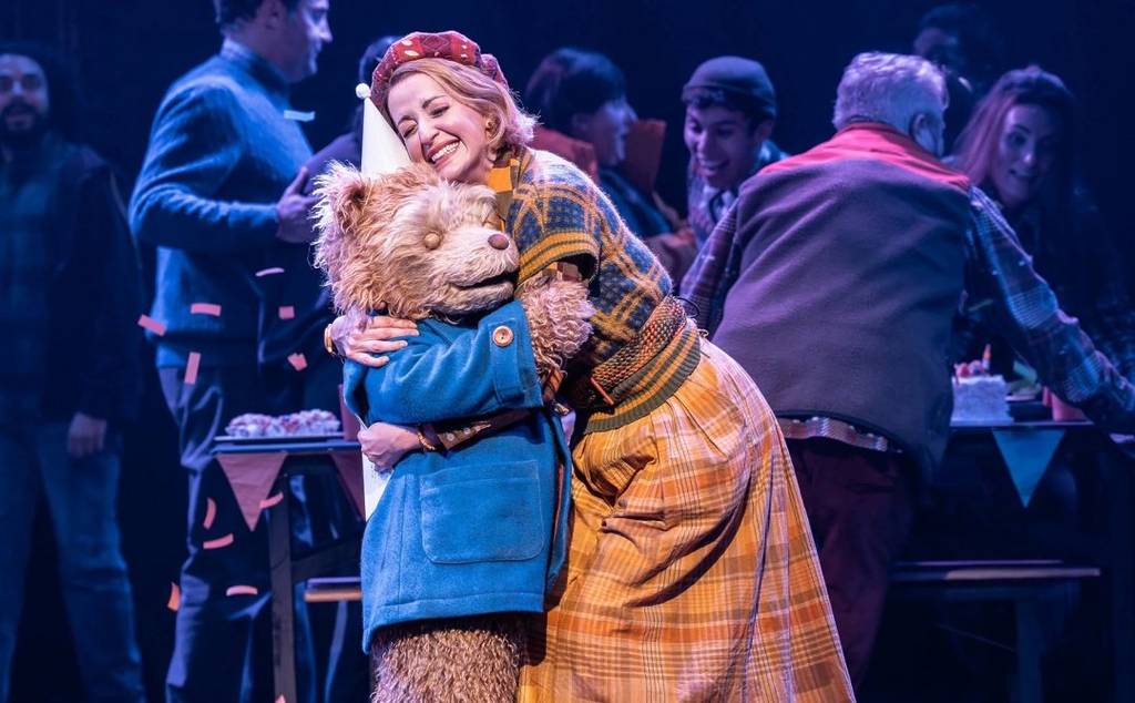 el oso Paddington en el escenario de Paddington: el musical, abrazando a alguien mientras lleva un gorro de fiesta