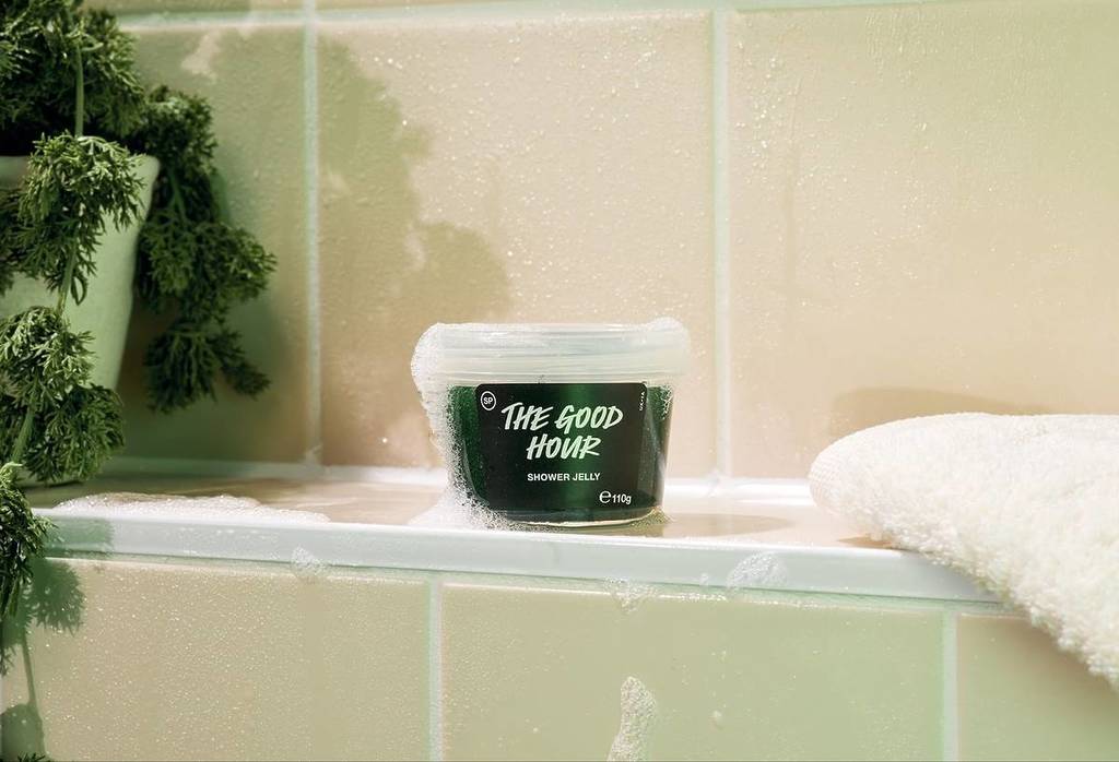 un bote de gel de ducha «The Good Hour» de LUSH —un producto de color verde oscuro en un bote de plástico— sobre una repisa de la ducha salpicada de espuma de jabón