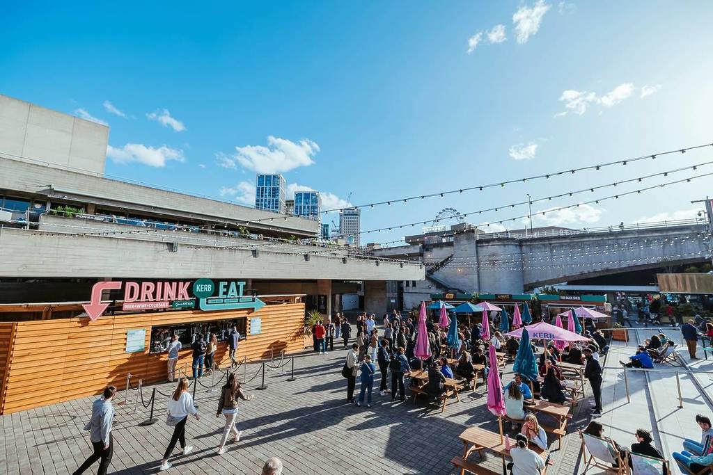 persone sedute ai tavoli sotto ombrelloni dai colori vivaci, accanto a un grande stand di cibo e bevande