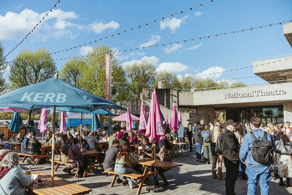 Menschen saßen an Tischen unter bunten Sonnenschirmen und schlenderten durch die Lebensmittelverkäufer, an einem sonnigen Tag auf dem Lebensmittelmarkt am Straßenrand des Nationaltheaters