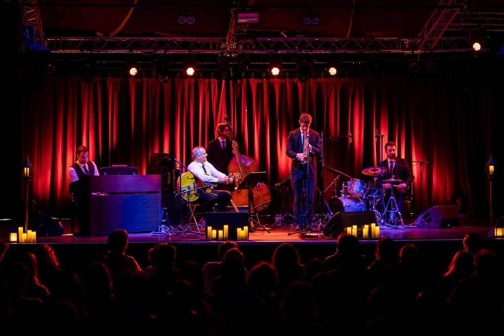 Een jazzband met vijf leden speelt een set op het podium in een sfeervol verlichte omgeving met rode gordijnen achter hen.