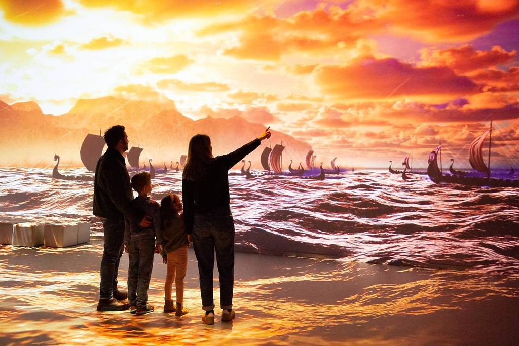 a family of four look at a giant projection of viking longships navigating rough seas in front of a sunset