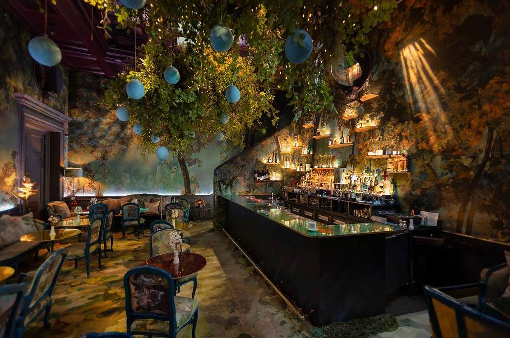 The interiors of the restaurant sketch transformed into the Hundred Acre Wood from Winnie the Pooh, with leafy branches sprouting across the ceiling with blue balloons hanging down and forest illustrated wallpaper bon the walls