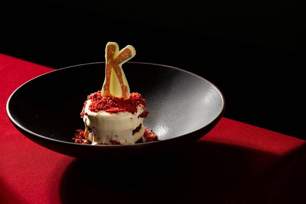 Een zwart bord met een wit, rond dessert met rode versieringen en het Kinky Boots "K"-logo er bovenop.