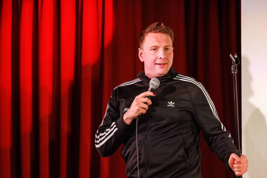 komiek Joe Lycett treedt op voor een rood fluwelen gordijn, met een microfoon in zijn hand