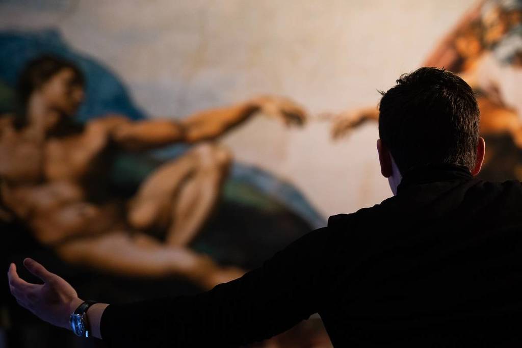 un hombre de pie con los brazos extendidos mientras observa una gran reproducción de la escena de La creación de Adán en la pintura de la Capilla Sixtina de Miguel Ángel