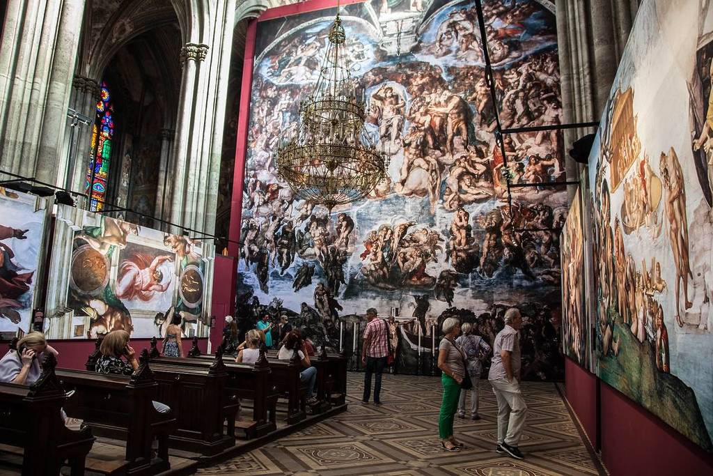 la gente pasea admirando reproducciones gigantes de la pintura de la Capilla Sixtina de Miguel Ángel, expuestas en una iglesia gótica