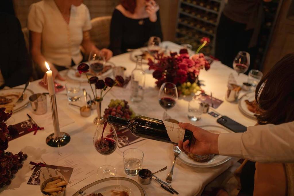 vino servido en una copa sobre una mesa con mantel blanco, llena de copas de vino y platos de comida, alrededor de la cual la gente se lo pasa en grande disfrutando de una buena comida