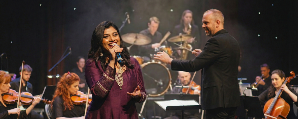 Priyani Vani Panditt sings into a microphone while a conductor and orchestra perform on a dark, smoky stage.