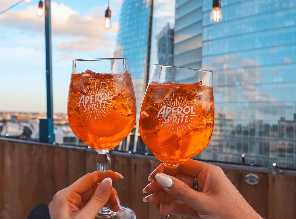 two hands cheers aperol spritz on a london rooftop with a skyscraper in the background