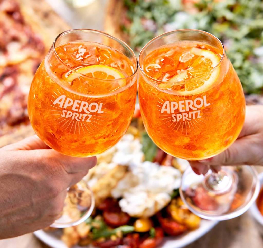 two aperol spritz drinks cheers over a table full of pizza