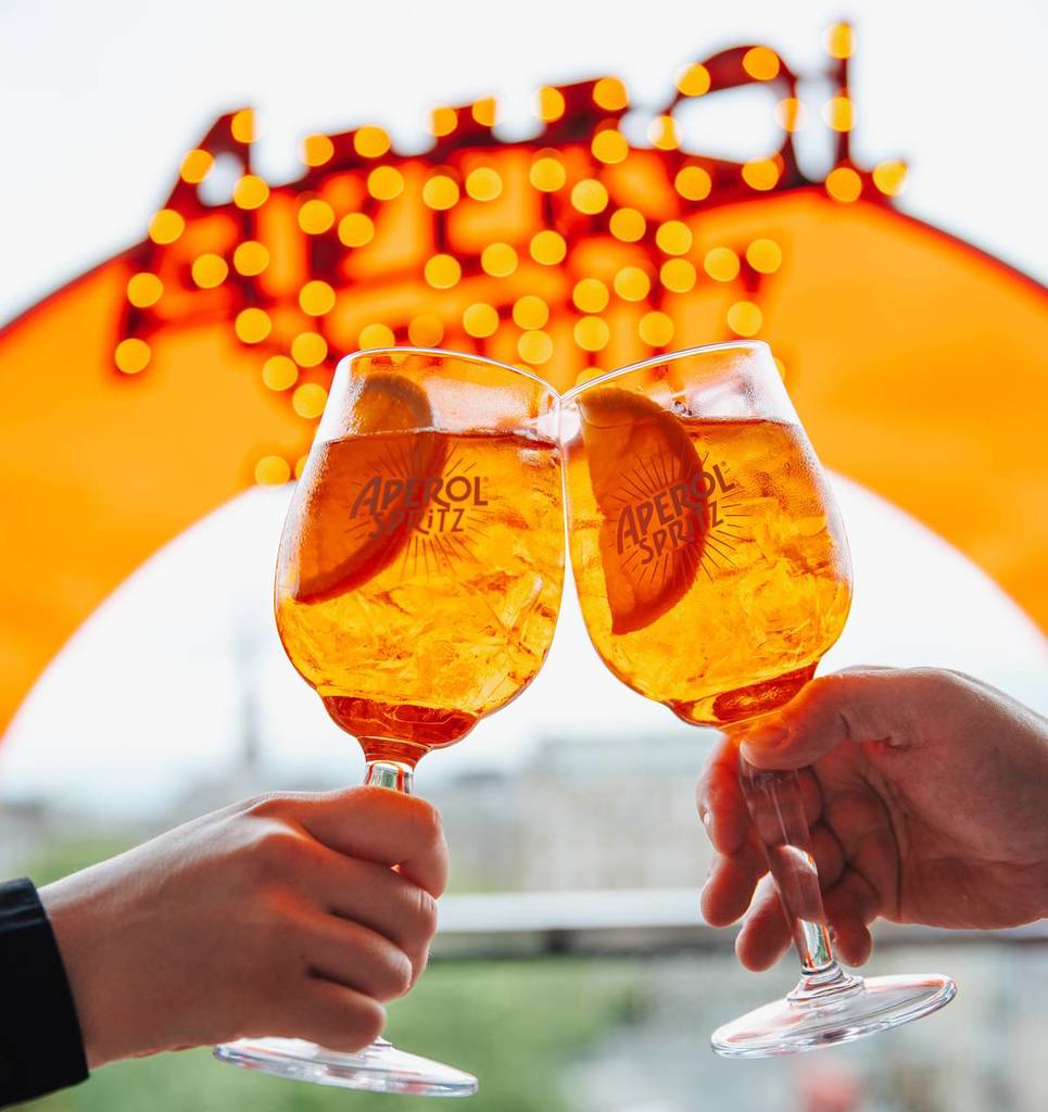 aperol spritz cheers on an outdoor terrace in front of an orange arch with the aperol logo in lights