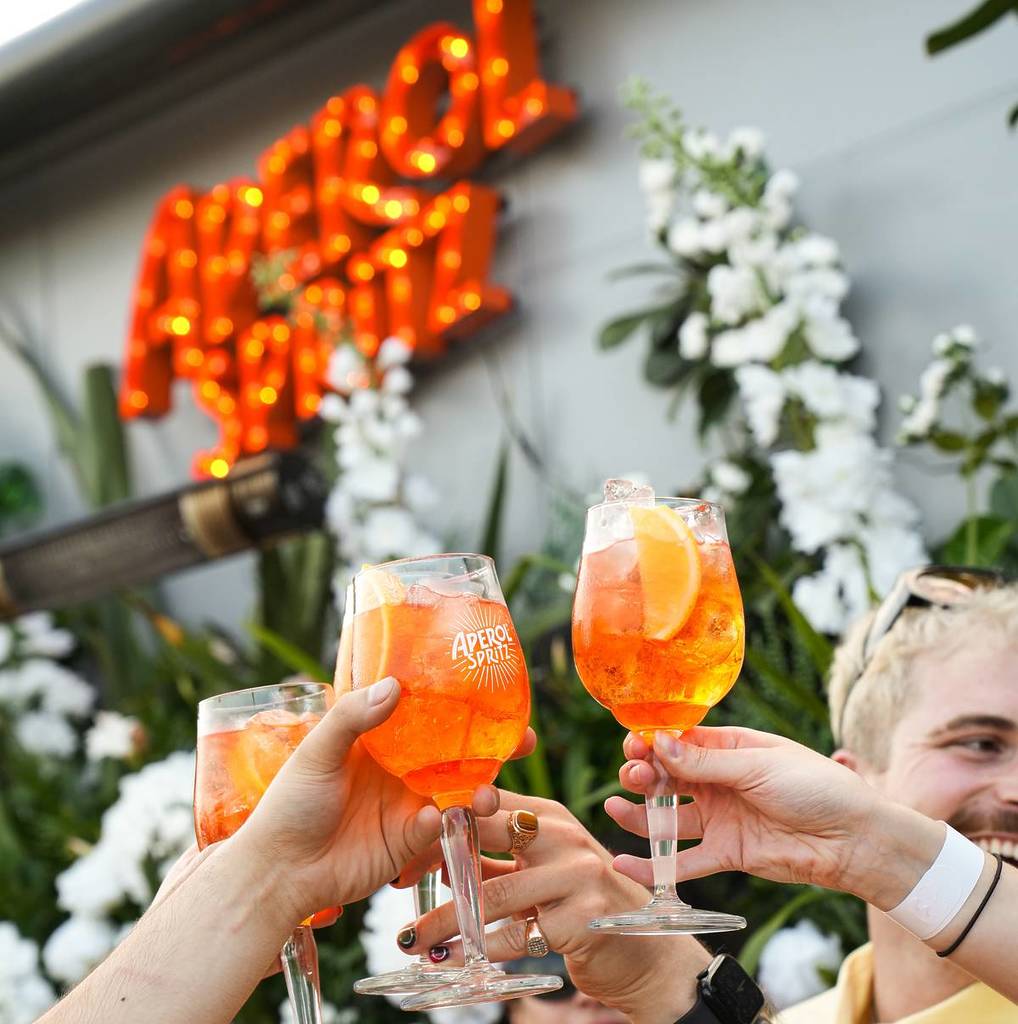 aperol spritz cheers in front of flowers with an aperol spritz sign in the background
