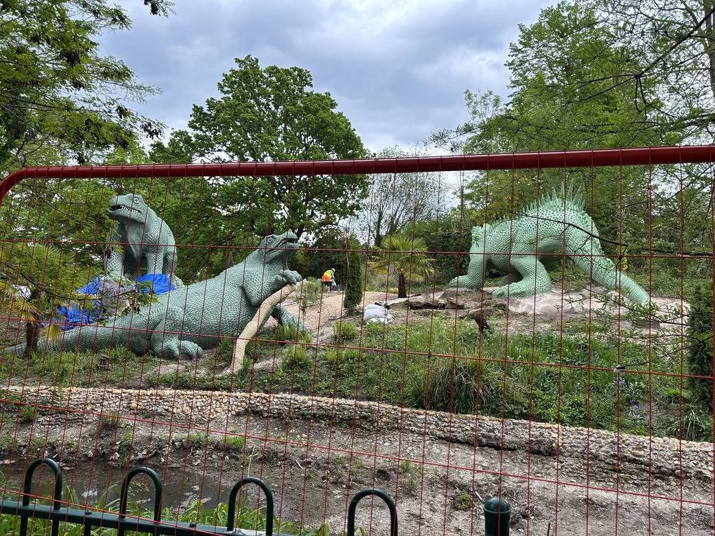 Grüne Skulpturen von Dinosauriern, wie sie hinter einem Bauzaun zu sehen sind, während sie restauriert werden