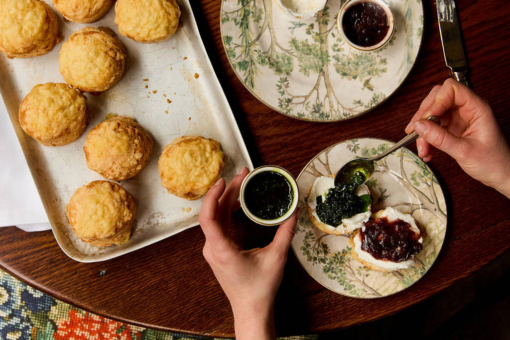 quelqu'un qui étale de la confiture sur un scone au fromage recouvert de caillé de chèvre, à côté d'un plateau de scones au fromage