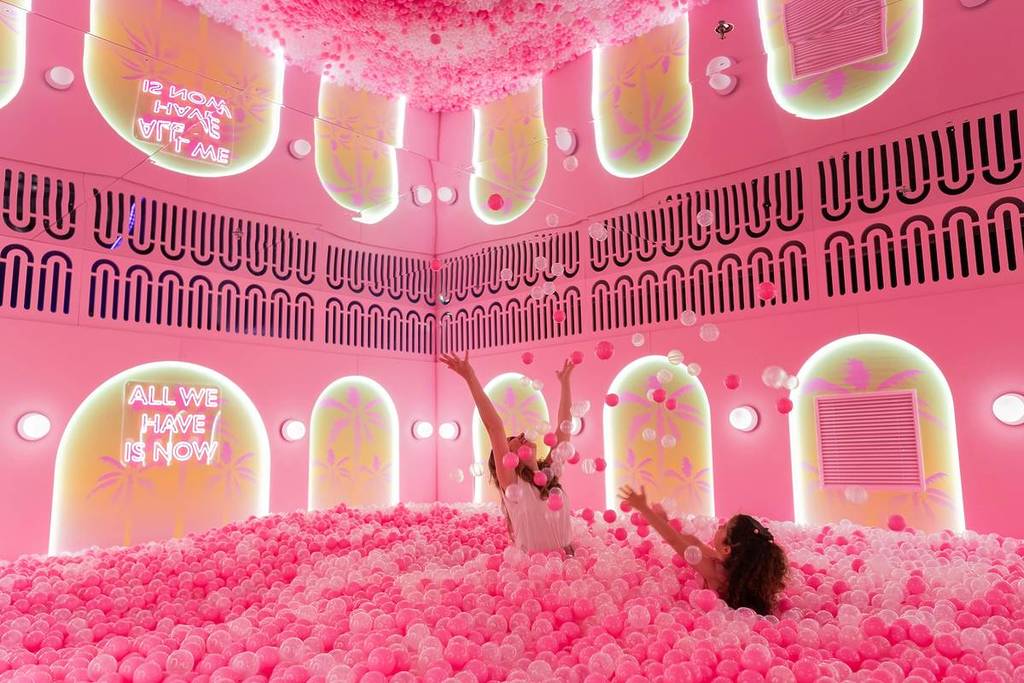 duas mulheres atiram bolas para o ar e riem-se numa piscina de bolas rosa vibrante com um teto espelhado, arcos luminosos na parede e letras de néon