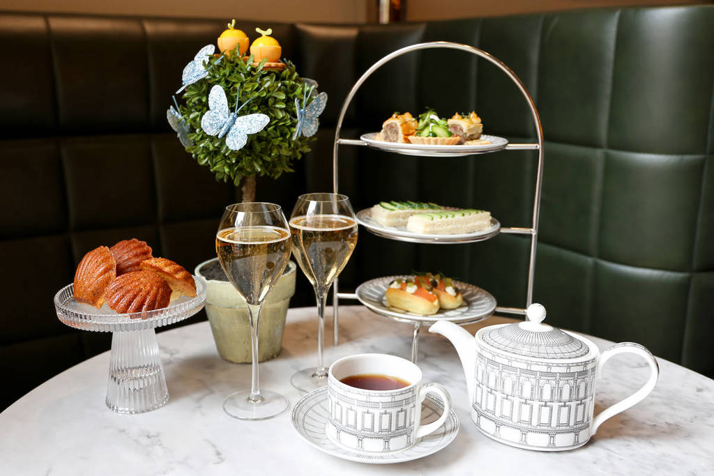 een afternoon tea-buffet met een etagère vol sandwiches en hartige hapjes, kopjes thee, glazen champagne, een schaal madeleines en een mandje vol eetbare lekkernijen