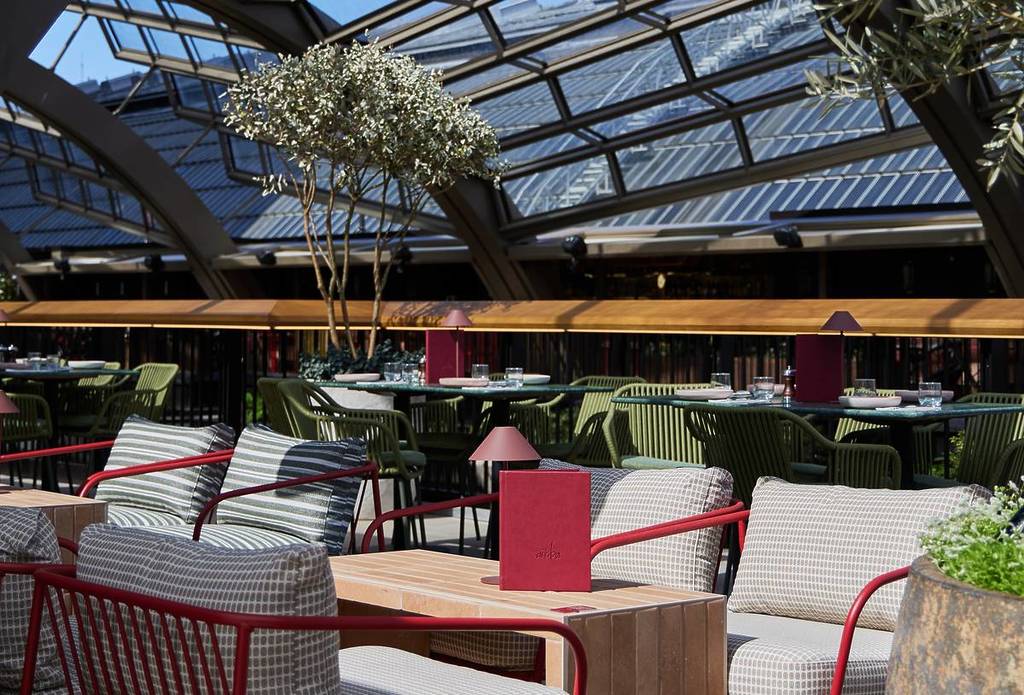 Una vista del Bar Arriba, un nuovo bar sul tetto a Olympia, con una disposizione semi-all’aperto caratterizzata da sedie rosse e verdi e tavoli da terrazza sotto un tetto in vetro piastrellato