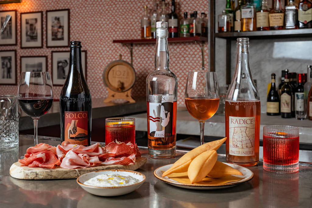 uma variedade de enchidos, queijos e petiscos fritos, acompanhados por garrafas de lambrusco
