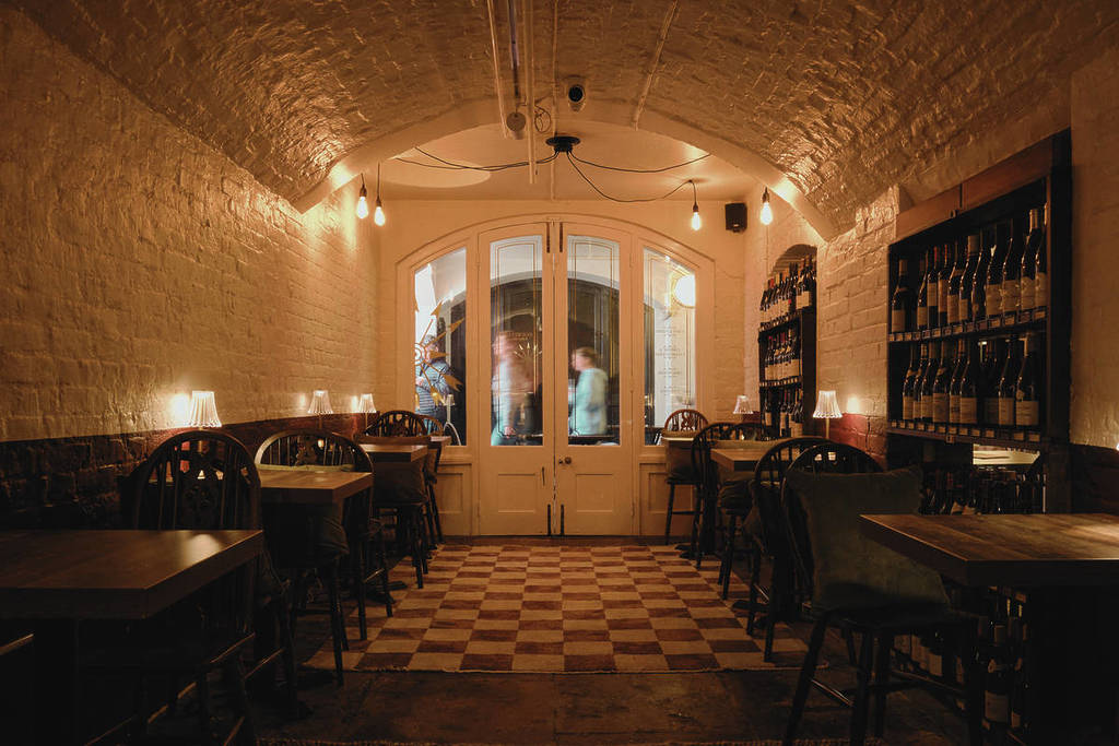 An dimly lit, rustic wine cellar interior with a vaulted ceiling, vintage-style tables and chairs, and wine shelves along the walls
