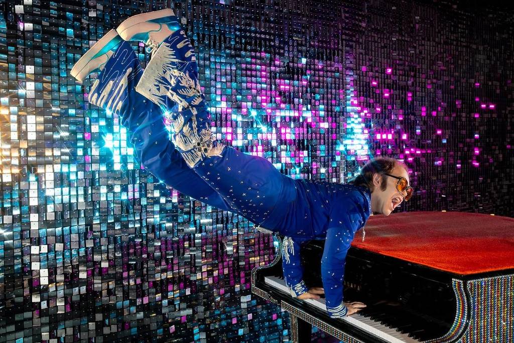een wassen beeld van Elton John in een glinsterende blauwe jumpsuit en een oranje bril die zijn iconische pianosprong maakt