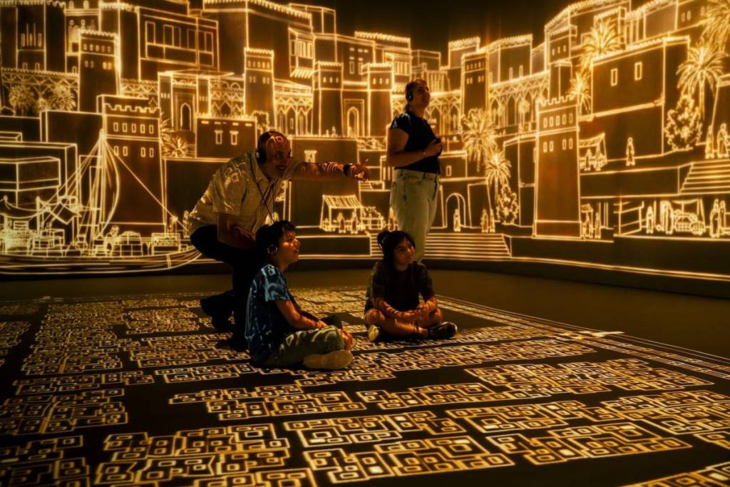 una famiglia seduta in una sala con le audioguide indosso, mentre osserva le proiezioni dorate di una città antica