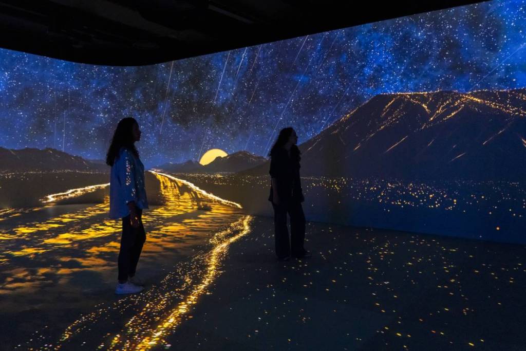 two women stand in a room with a projection of a golden river running into the mountains and a sunset on the horizon