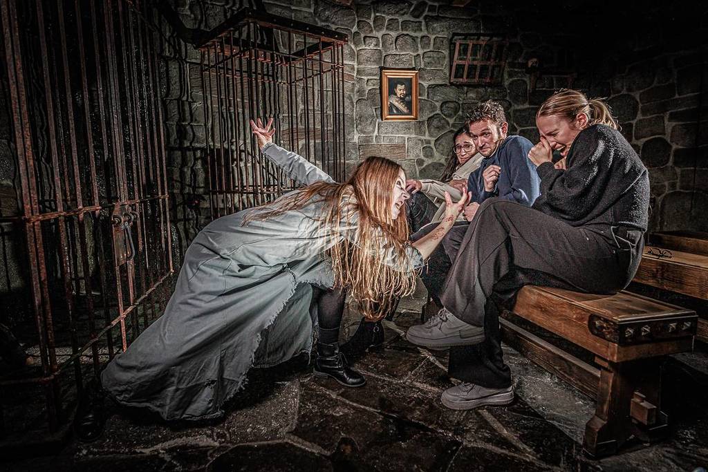 An actress dressed in a grey gown with long lank hair reaches out aggressively towards three people sitting in the audience in a prison-style room at the London Dungeon