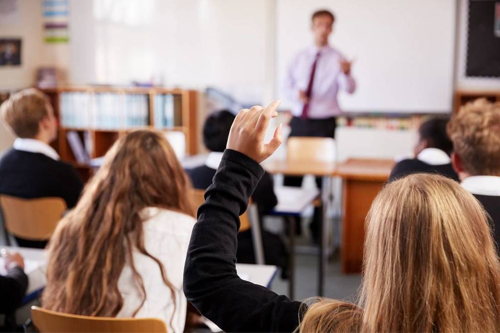 Ein Klassenzimmer von hinten, ein Mädchen hebt die Hand, andere Schüler sind an ihren Schreibtischen zu sehen und im Hintergrund eine Lehrerin