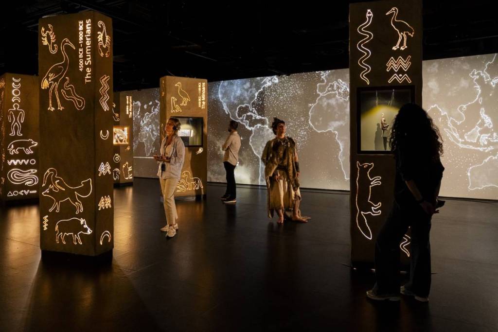 la gente pasea por una sala llena de columnas grabadas con símbolos luminosos de civilizaciones antiguas en la exposición «The Timewalk»