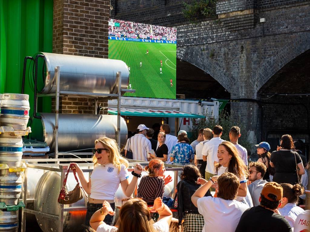 gente con camisetas de Inglaterra animando y paseándose mientras ven el partido de Inglaterra en una pantalla gigante en un patio